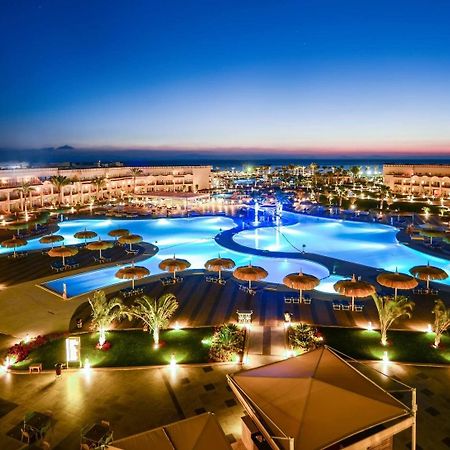 Pickalbatros Royal Moderna Sharm "Aqua Park" Hotel Exterior photo The photo showcases an elegant resort area at dusk. It features a large swimming pool, which is beautifully illuminated with blue lights, creating a serene and inviting atmosphere. Surrounding the pool are palm trees and umbrellas, enhancing the trop