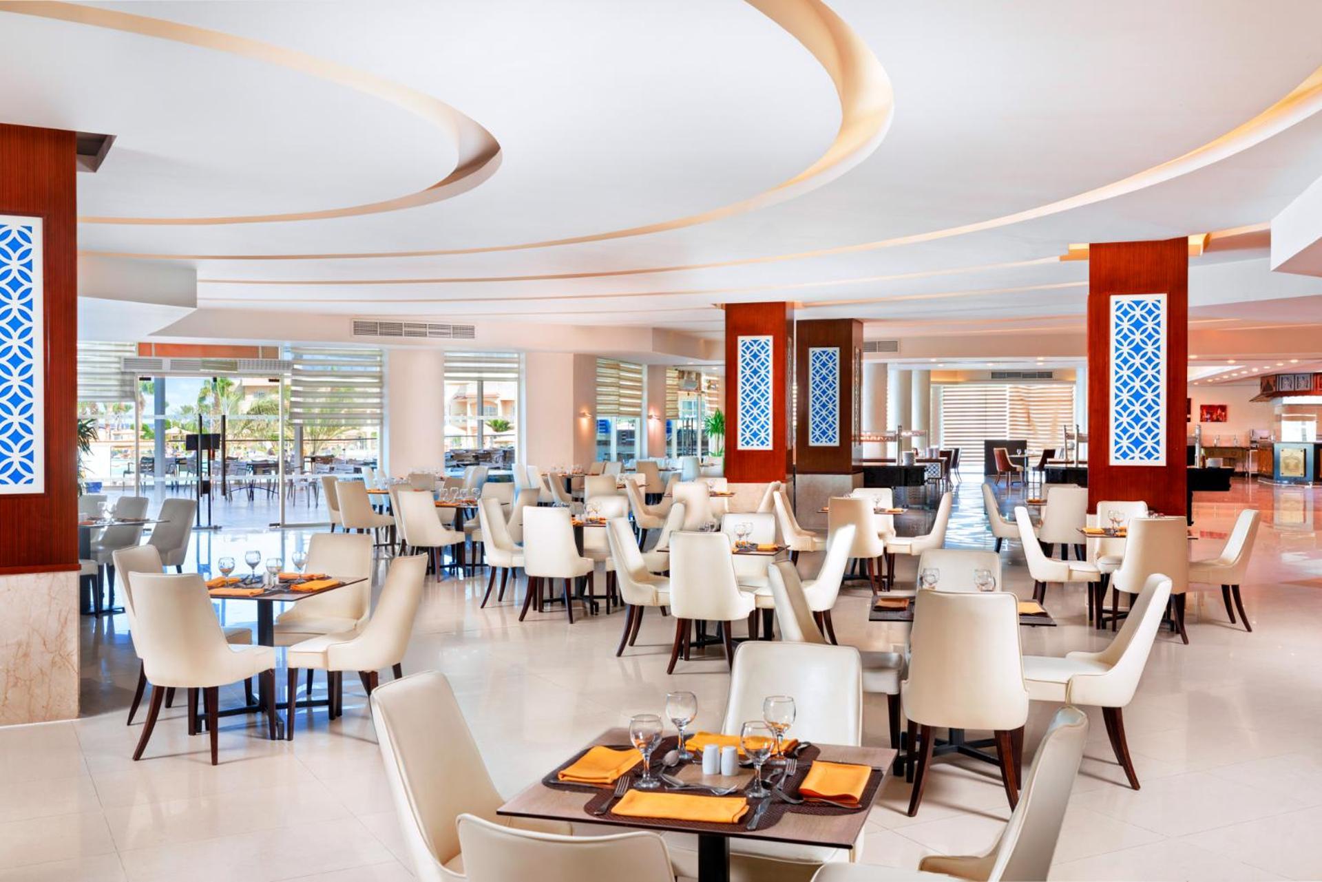 Pickalbatros Royal Moderna Sharm "Aqua Park" Hotel Exterior photo The photo shows a modern restaurant interior. The design features light, neutral colors with elegant white chairs and wooden tables. The layout includes several dining tables set for customers, creating a spacious and inviting atmosphere. There are d