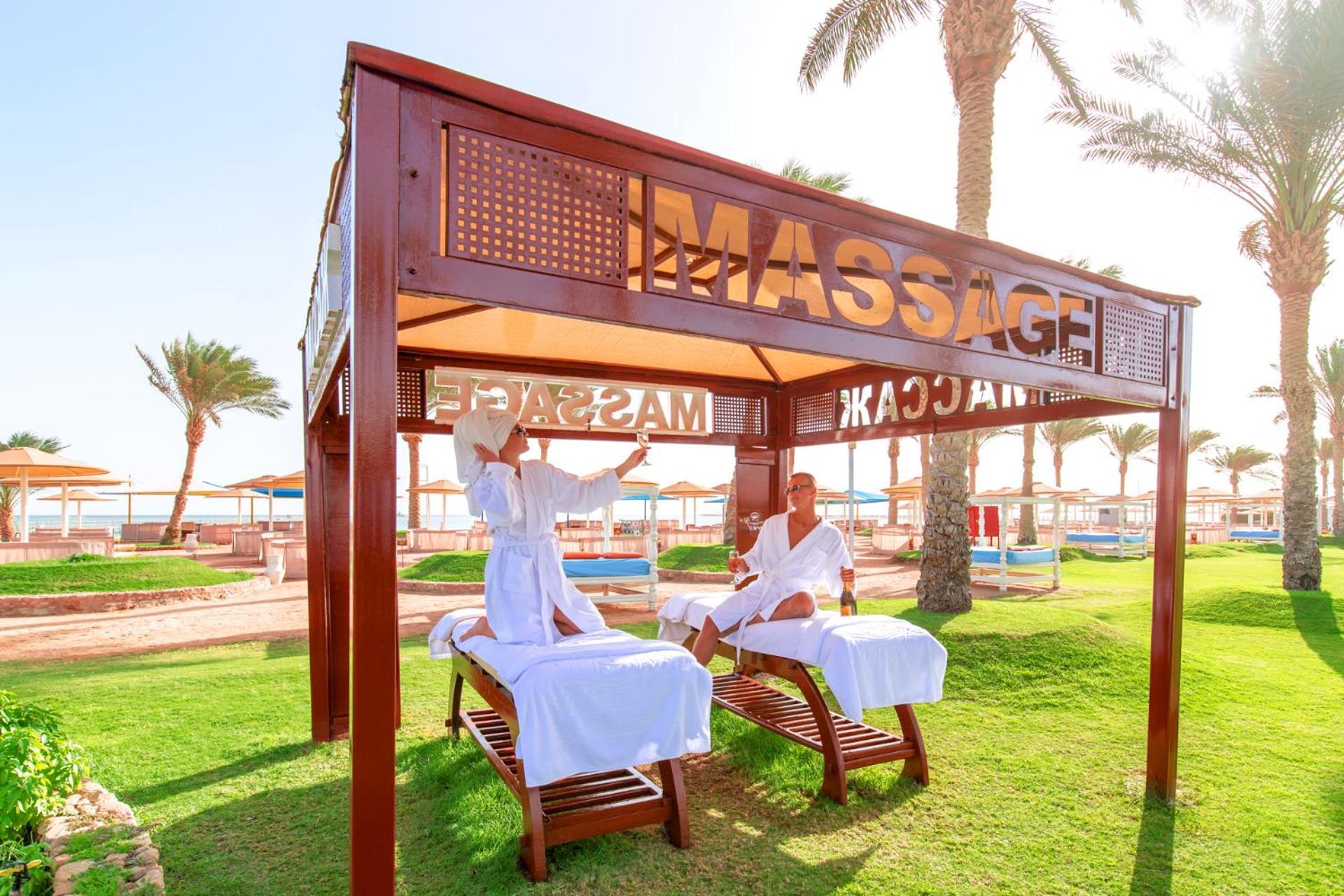 Pickalbatros Royal Moderna Sharm "Aqua Park" Hotel Exterior photo The photo shows a massage area set up in a sunny outdoor environment. There are two massage tables under a wooden canopy with the word "MASSAGE" prominently displayed. Two individuals, dressed in white robes, appear to be engaged in a massage session