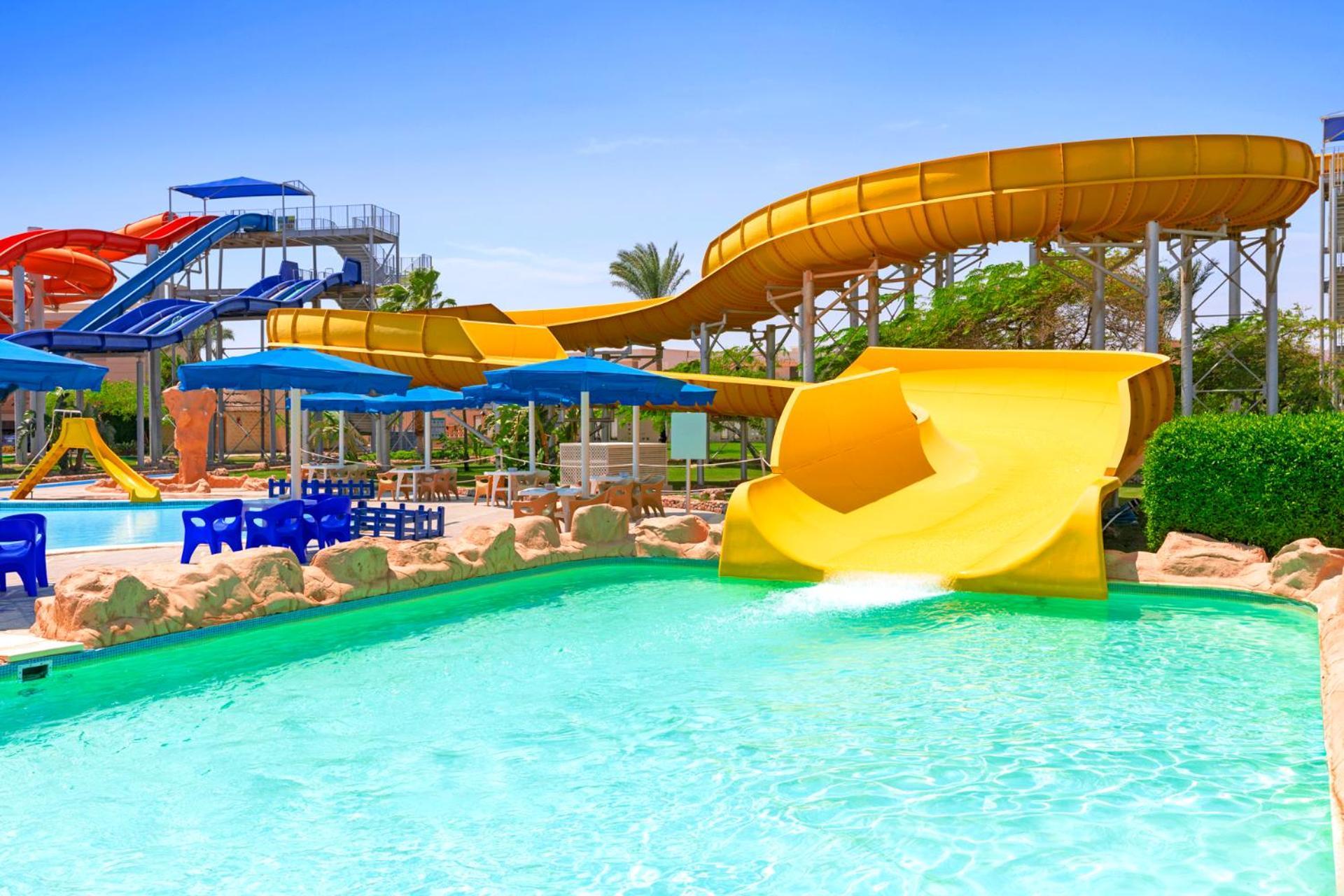 Pickalbatros Royal Moderna Sharm "Aqua Park" Hotel Exterior photo The image depicts a vibrant water park setting. In the foreground, there is a clear blue swimming pool with a yellow water slide curving into it. Next to the yellow slide, there's another water slide that is orange and red, elevating above the pool a
