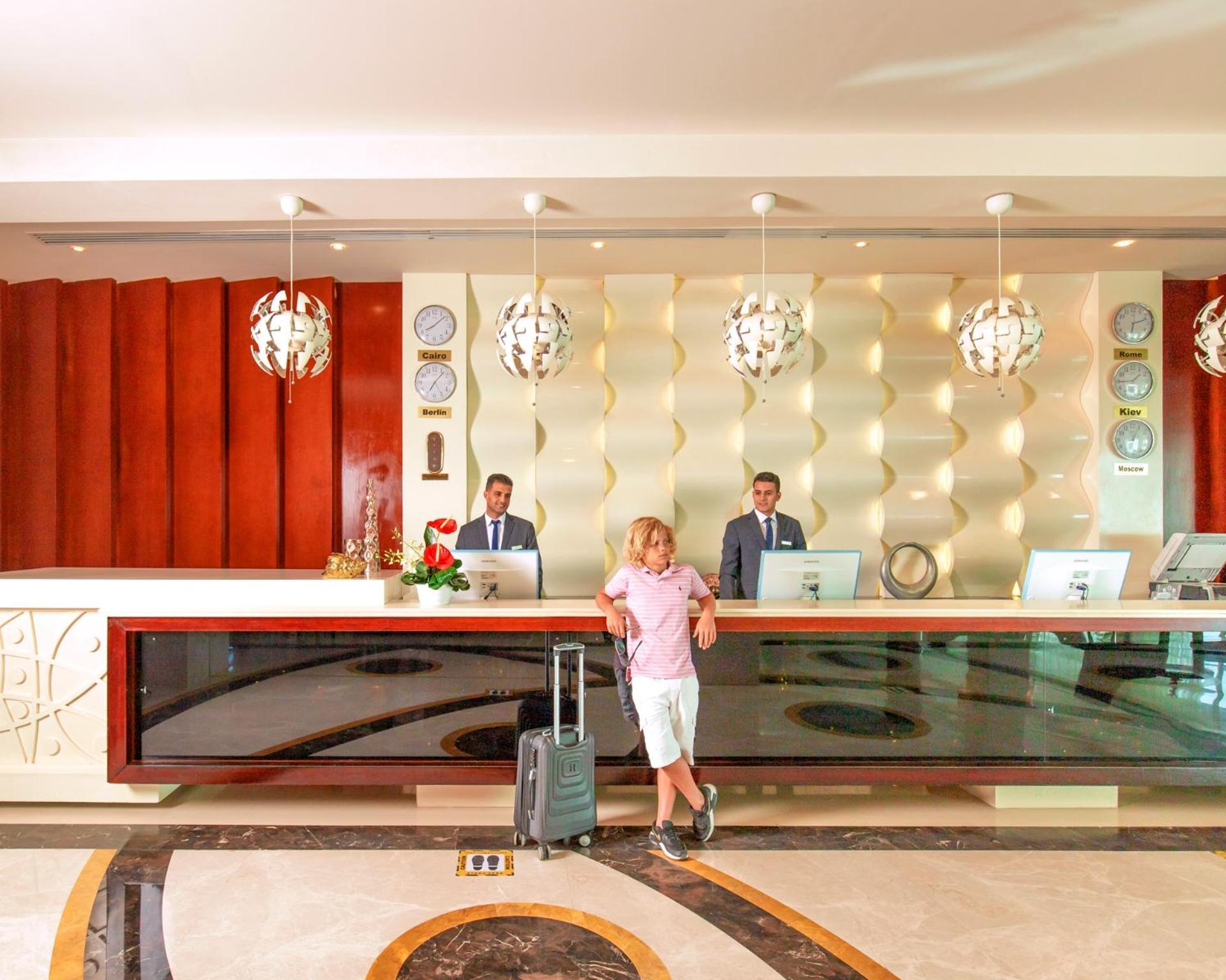 Pickalbatros Royal Moderna Sharm "Aqua Park" Hotel Exterior photo The photo depicts a hotel reception area. At the front desk, two staff members in suits are standing, presumably ready to assist guests. A man in casual attire, holding a suitcase, is standing in front of the desk. The reception has a modern design w