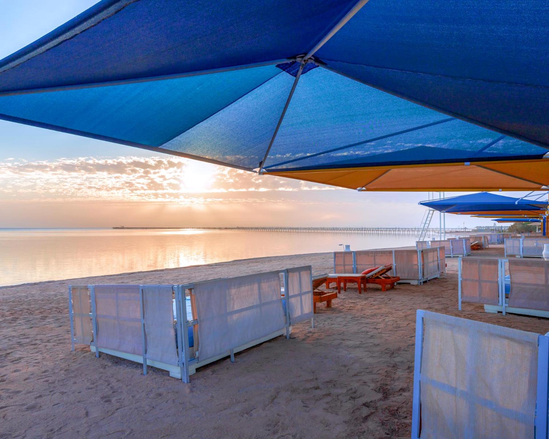 Pickalbatros Royal Moderna Sharm "Aqua Park" Hotel Exterior photo The photo shows a beach scene during sunset. There are colorful umbrellas in various shades of blue and orange set up over sandy areas. In the foreground, there are some wooden lounge chairs, and behind them, partition-like structures or beach cabana