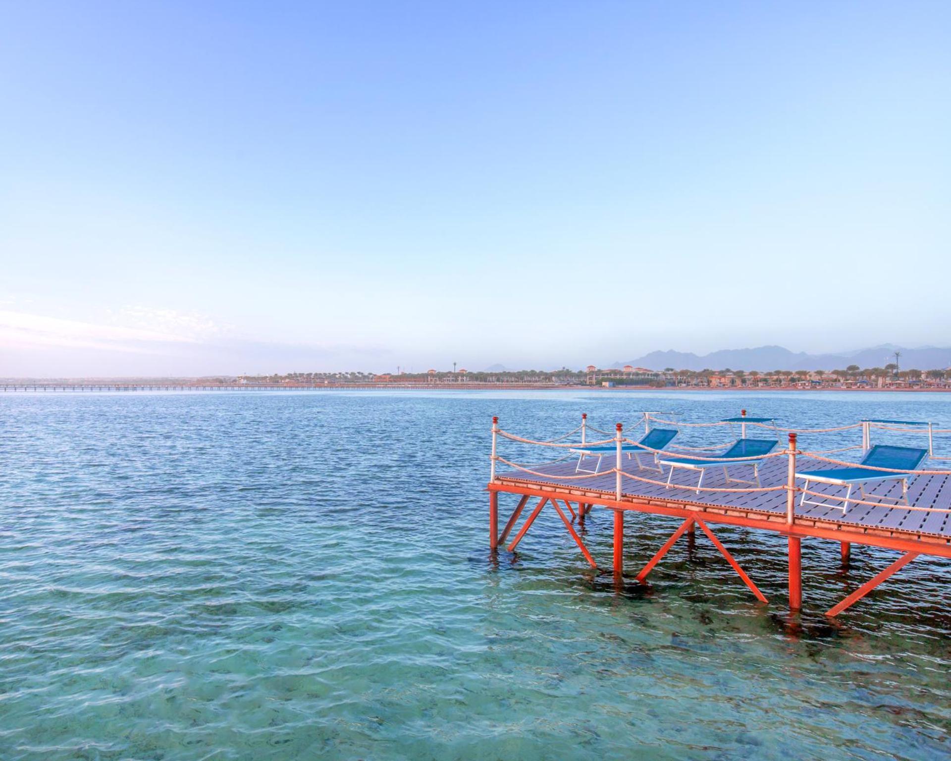 Pickalbatros Royal Moderna Sharm "Aqua Park" Hotel Exterior photo The photo shows a peaceful scene featuring a wooden dock extending into calm water. The dock is painted orange and has several blue lounge chairs placed on it, suggesting a relaxed atmosphere. In the background, there's a shoreline with some greenery