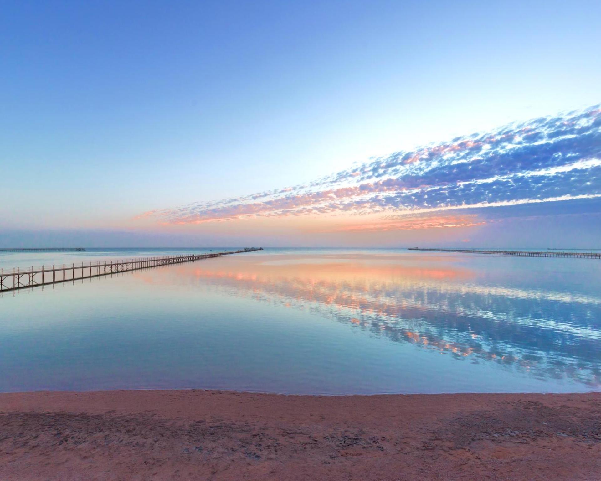 Pickalbatros Royal Moderna Sharm "Aqua Park" Hotel Exterior photo The photo depicts a serene coastal scene at dawn or dusk. There is a calm body of water that reflects the soft hues of the sky, which include shades of blue, pink, and orange. A long wooden pier extends into the water, adding depth to the composition