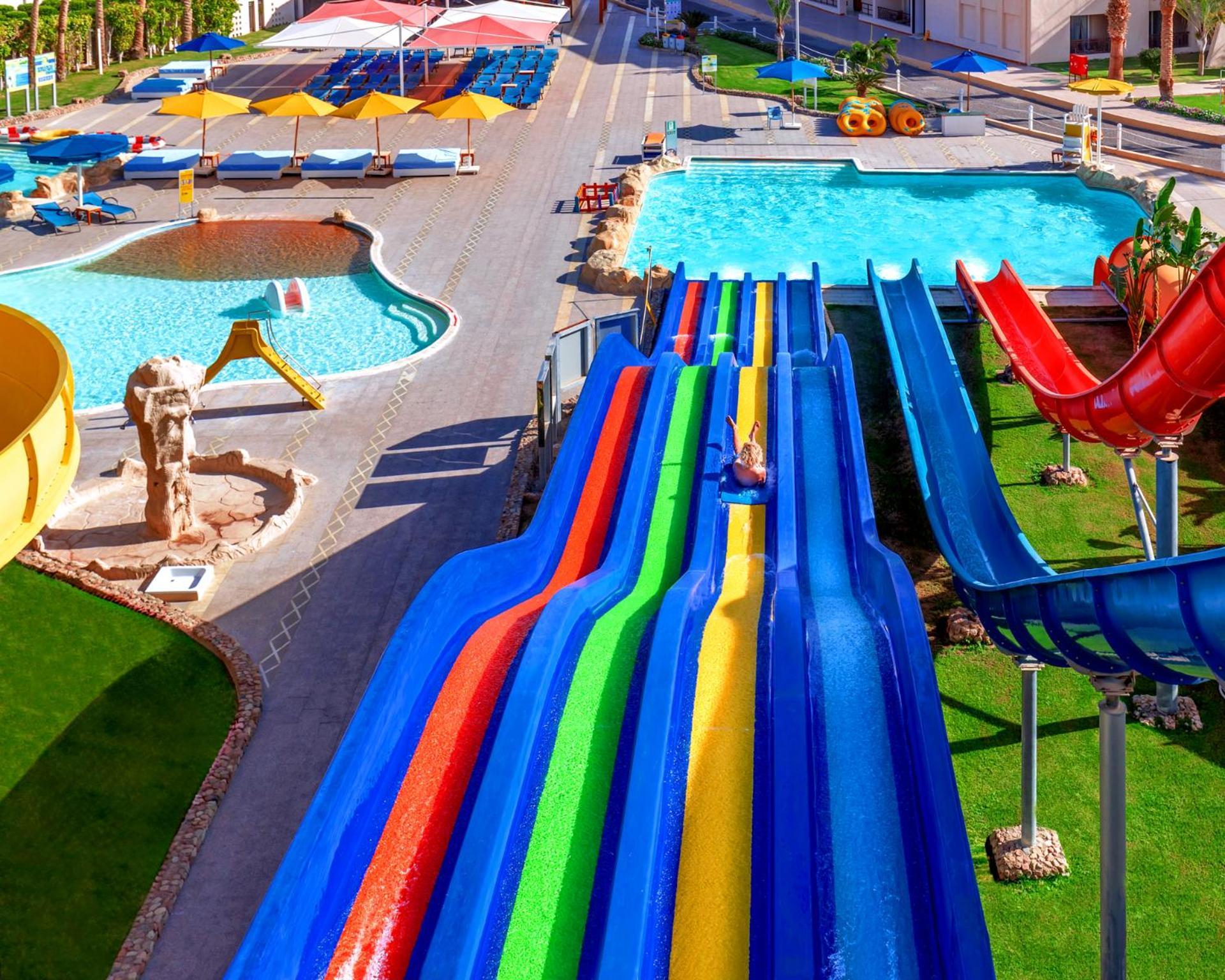Pickalbatros Royal Moderna Sharm "Aqua Park" Hotel Exterior photo The photo shows a vibrant water park featuring several large water slides in various colors, including red, blue, green, and yellow. There are swimming pools nearby, one of which has a clear blue appearance. The area is surrounded by green grass and 