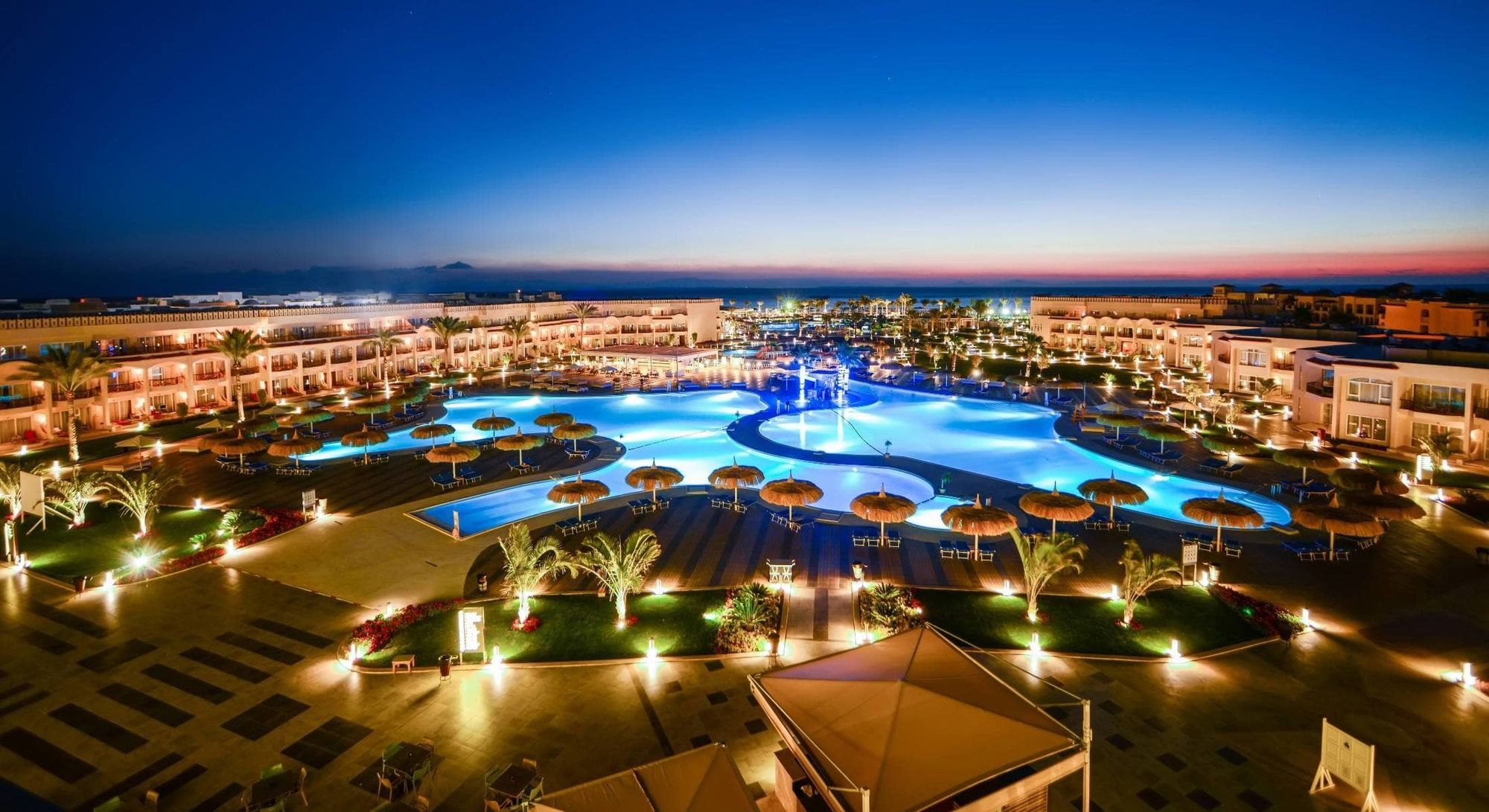 Pickalbatros Royal Moderna Sharm "Aqua Park" Hotel Exterior photo The photo showcases an elegant resort area at dusk. It features a large swimming pool, which is beautifully illuminated with blue lights, creating a serene and inviting atmosphere. Surrounding the pool are palm trees and umbrellas, enhancing the trop